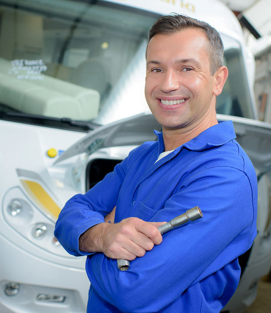 Smiling technician in blue uniform holding a wrench in front of an RV.